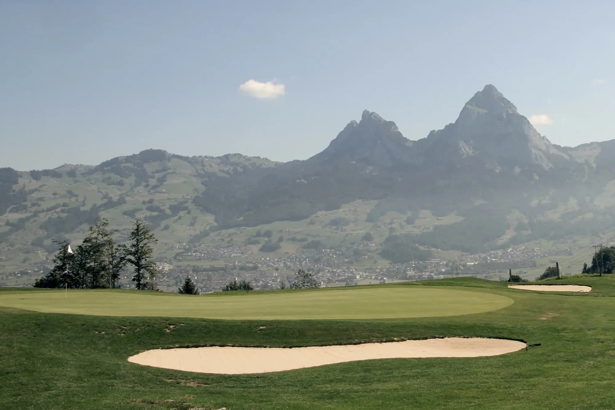 Golfgrün mit Bunker, dahinter die Mythen im Dunst.